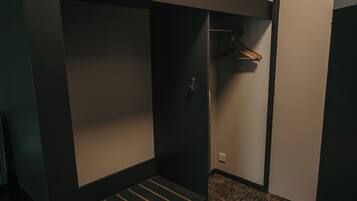 Um quarto pequeno e com pouca luz, com uma cama de solteiro, um ventilador de teto e um cabideiro.