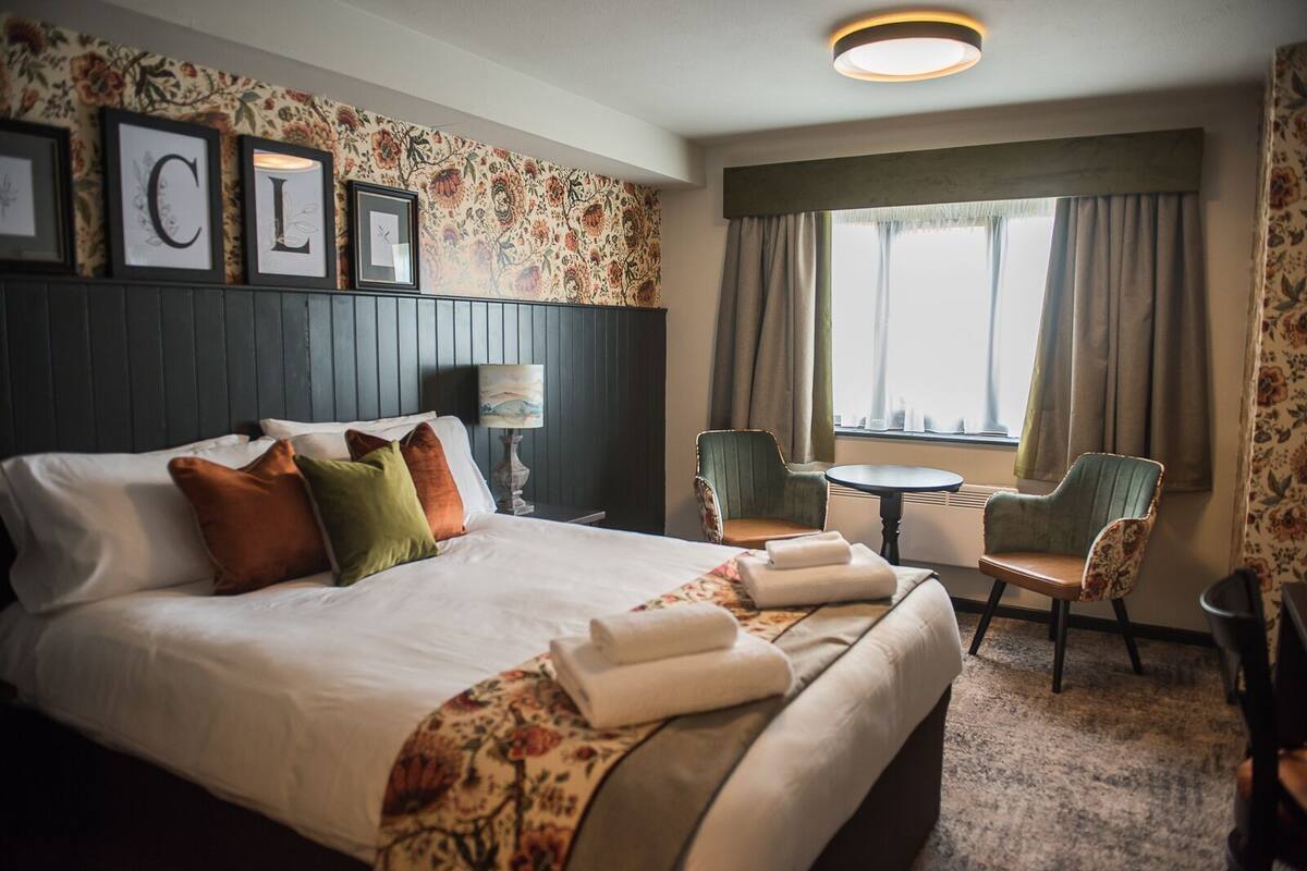 A hotel room with a bed, a chair, a small table, and a window with curtains.