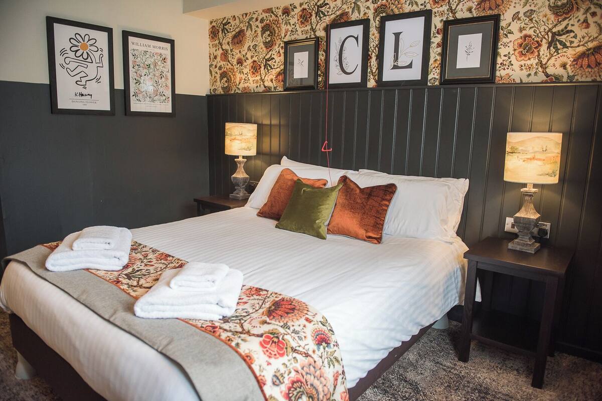 A neatly made bed with a floral bedspread, two pillows, and two folded towels. There are two bedside lamps and a framed artwork above the bed.