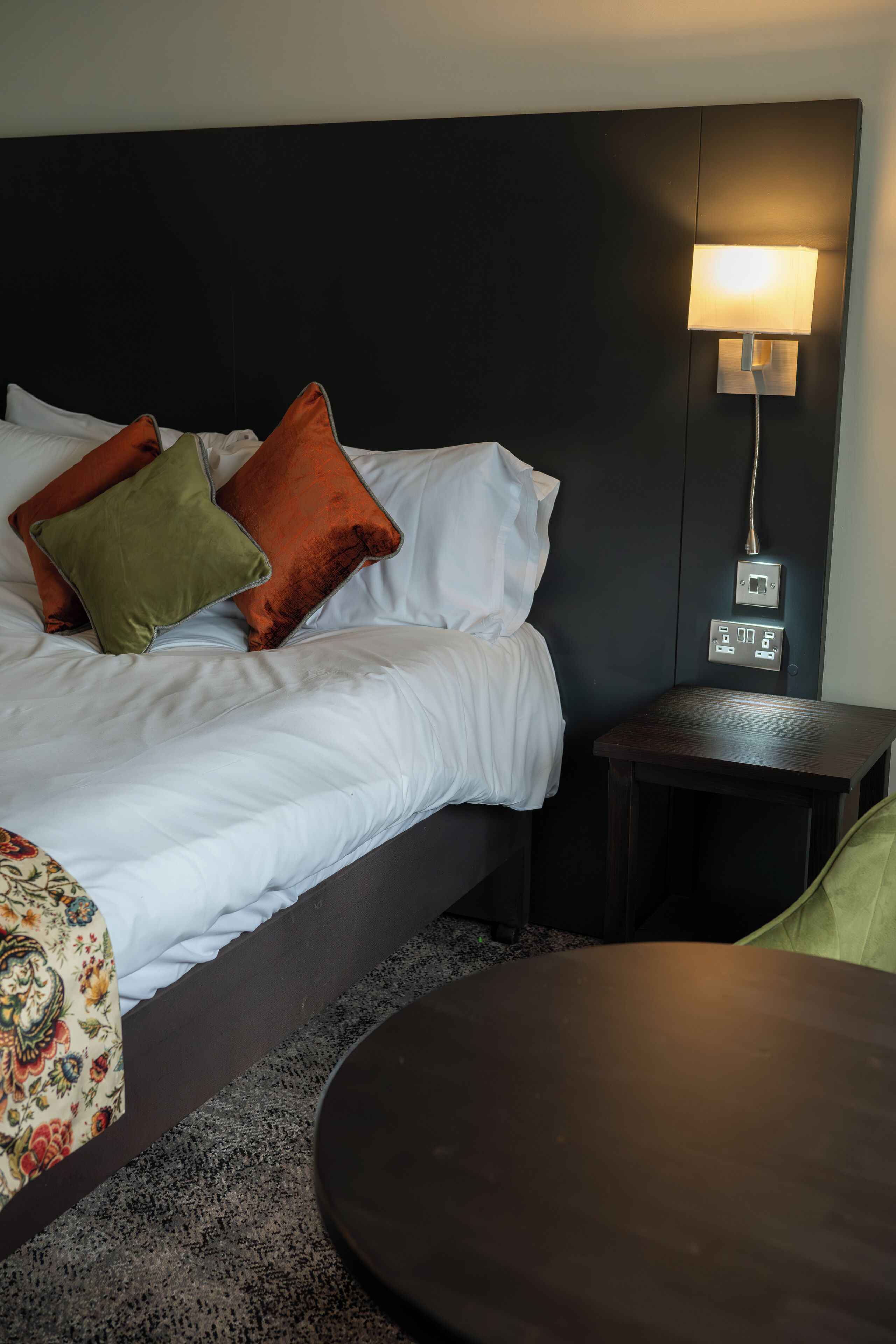 A hotel room with a bed, pillows, a bedside table, and a lamp.