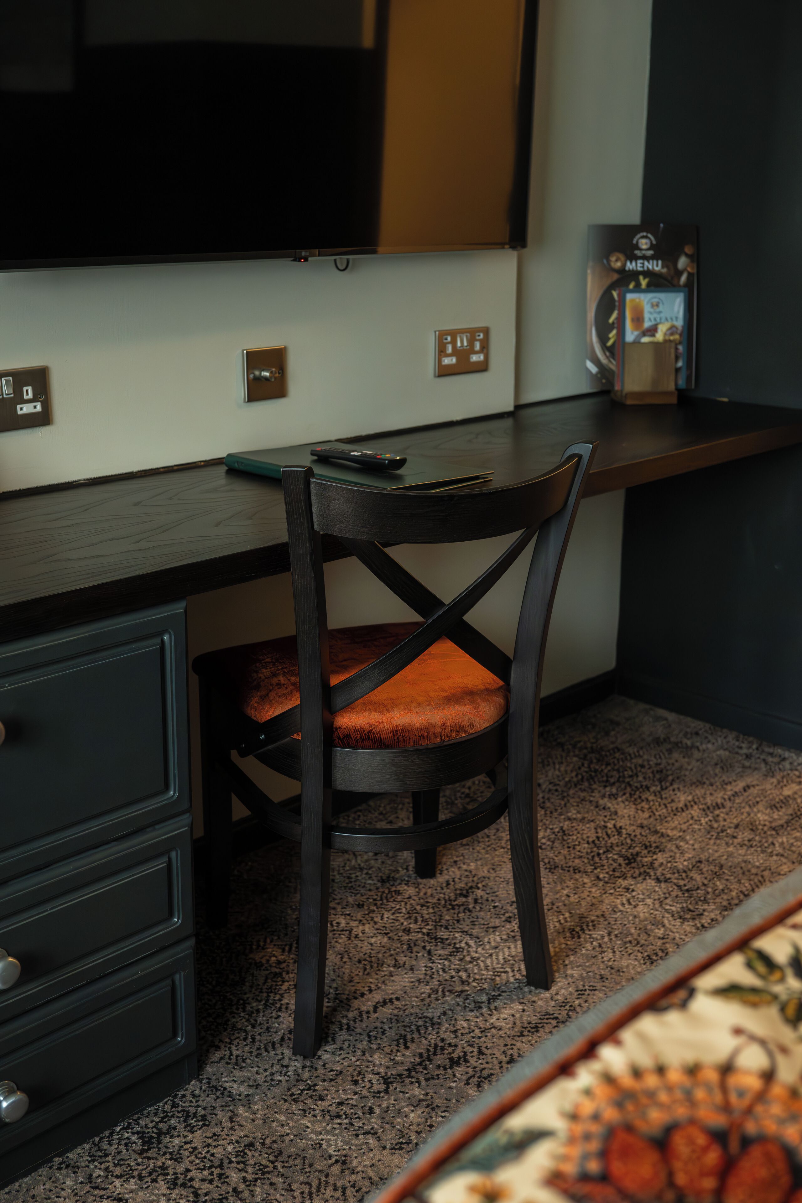 Ein Zimmer mit dunklem Holztisch, einem Stuhl mit gepolstertem Sitz, einem kleinen Tisch mit einer Uhr und einer Kommode.