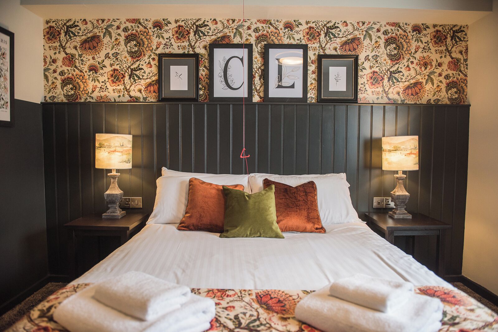 A neatly made bed with white linens and multiple pillows, flanked by two bedside tables with lamps, set against a wall with floral wallpaper and framed artwork.