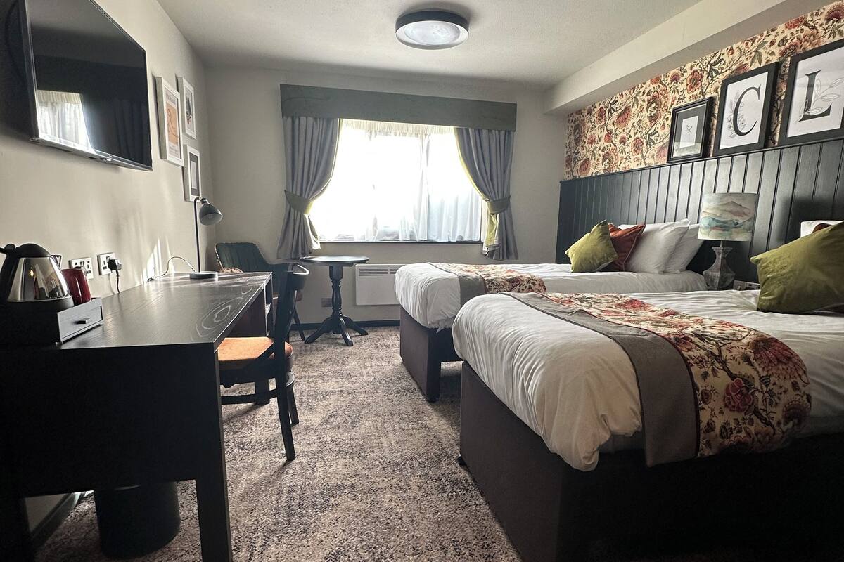 A hotel room with a bed, a desk, and a chair. There is a window with curtains, a flat-screen TV mounted on the wall, and a ceiling light.