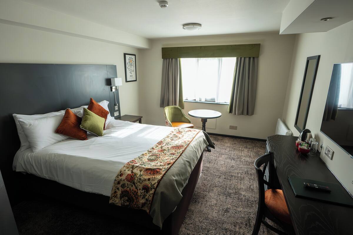 A hotel room with a bed, a desk, a chair, a round table, and a large window with curtains.