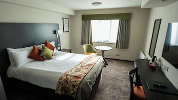 Habitación de hotel con cama, escritorio, silla, mesa redonda y un ventanal con cortinas.