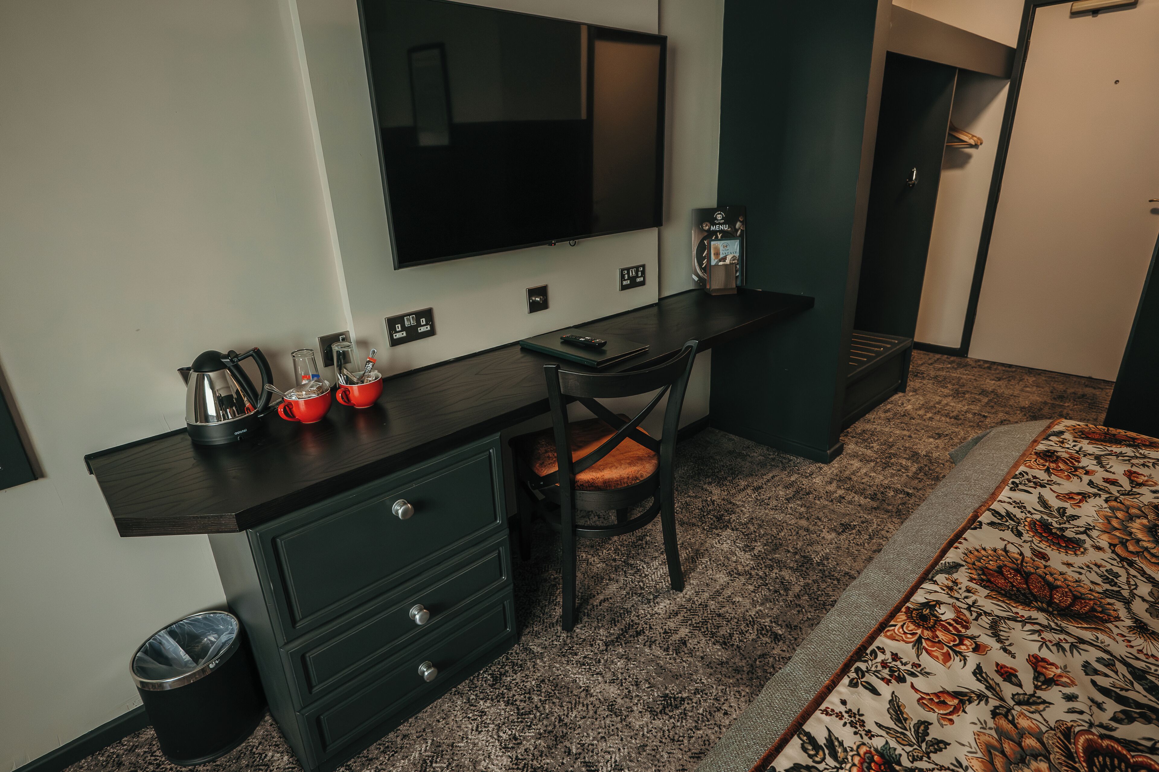 Une chambre d’hôtel de taille modeste, dotée d’un bureau en bois foncé, d’une seule chaise, d’un grand miroir, d’une télévision et d’une couvre-lit à motifs.
