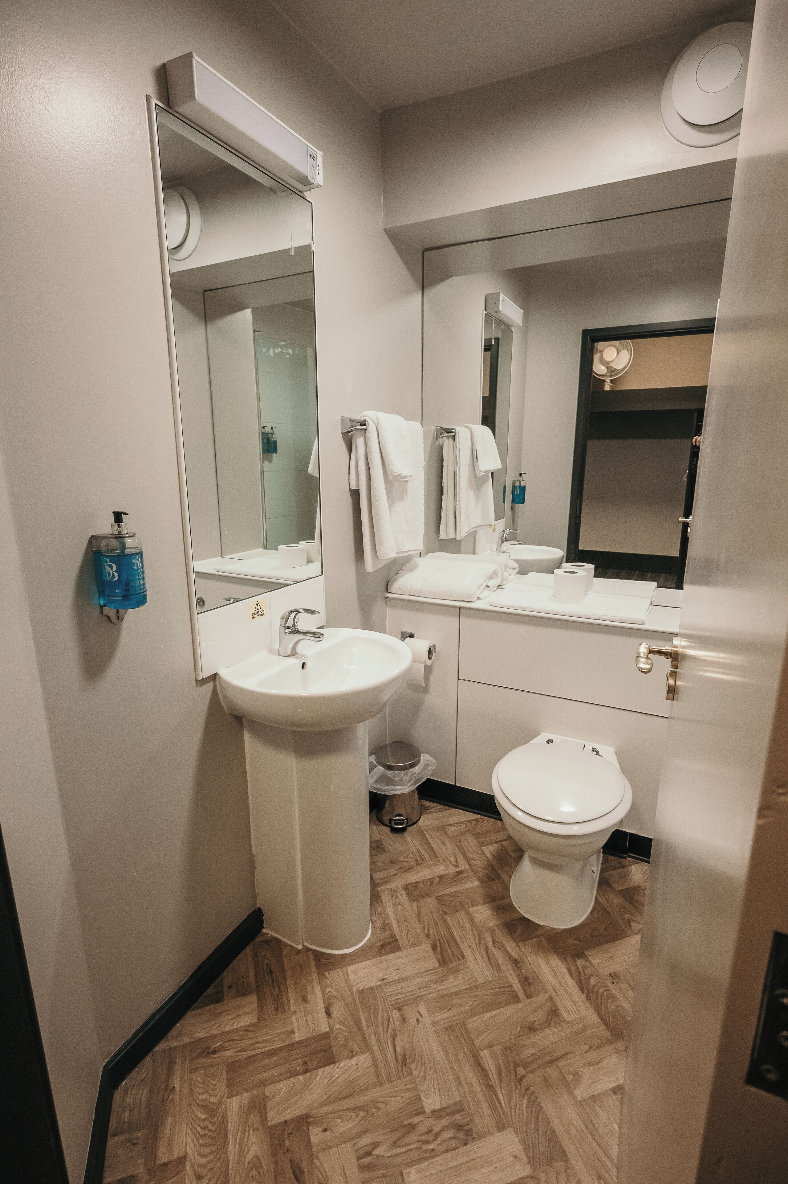 Casa de banho moderna com lavatório branco, espelho e sanita. Dispenser de sabonete azul na parede, e toalhas penduradas de forma organizada num suporte.