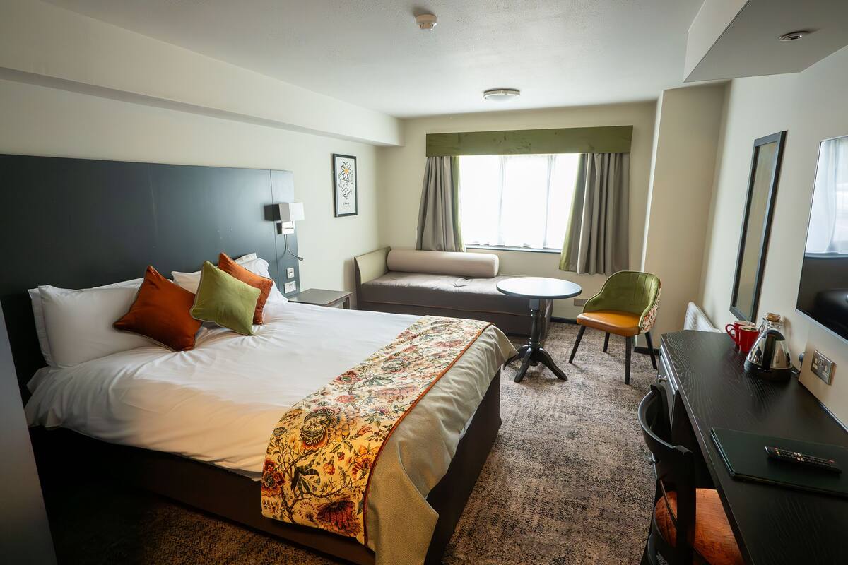 A hotel room with a bed, a desk, a chair, a small table, and a window with curtains.