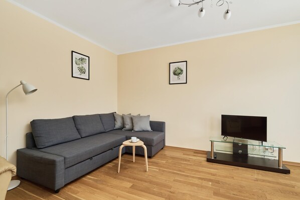 A modern living room with a grey sectional sofa, a small wooden side table, a glass TV stand, and two framed botanical prints on the walls.