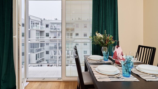 A dining area with a table set for four, a view of a residential building through a sliding glass door, and a window with blinds. - Spacious Apartment | Balcony, Parking, Workspace (Wroclaw)