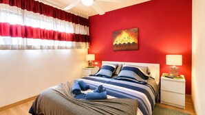 Ein Schlafzimmer mit einem Bett, zwei Nachttischen, einem Deckenventilator, einer roten Wand und einem Gemälde.