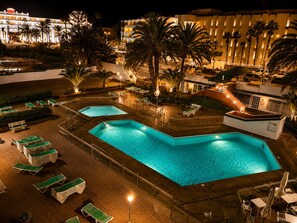 Nachtsicht eines Hotels mit zwei Schwimmbecken, Liegestühlen und Palmen.