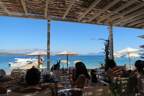 An outdoor seating area with tables and umbrellas overlooking a beach and the sea.