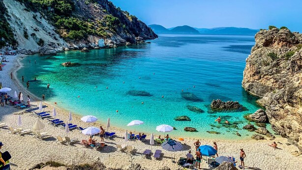 Une plage avec une eau turquoise et limpide, des falaises rocheuses et une rive sablonneuse agrémentée de transats et de parasols.