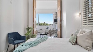 Ein Schlafzimmer mit einem großen Bett, einem blauen Sessel, Blick ins Freie und einem Balkon mit Tisch und Stühlen.