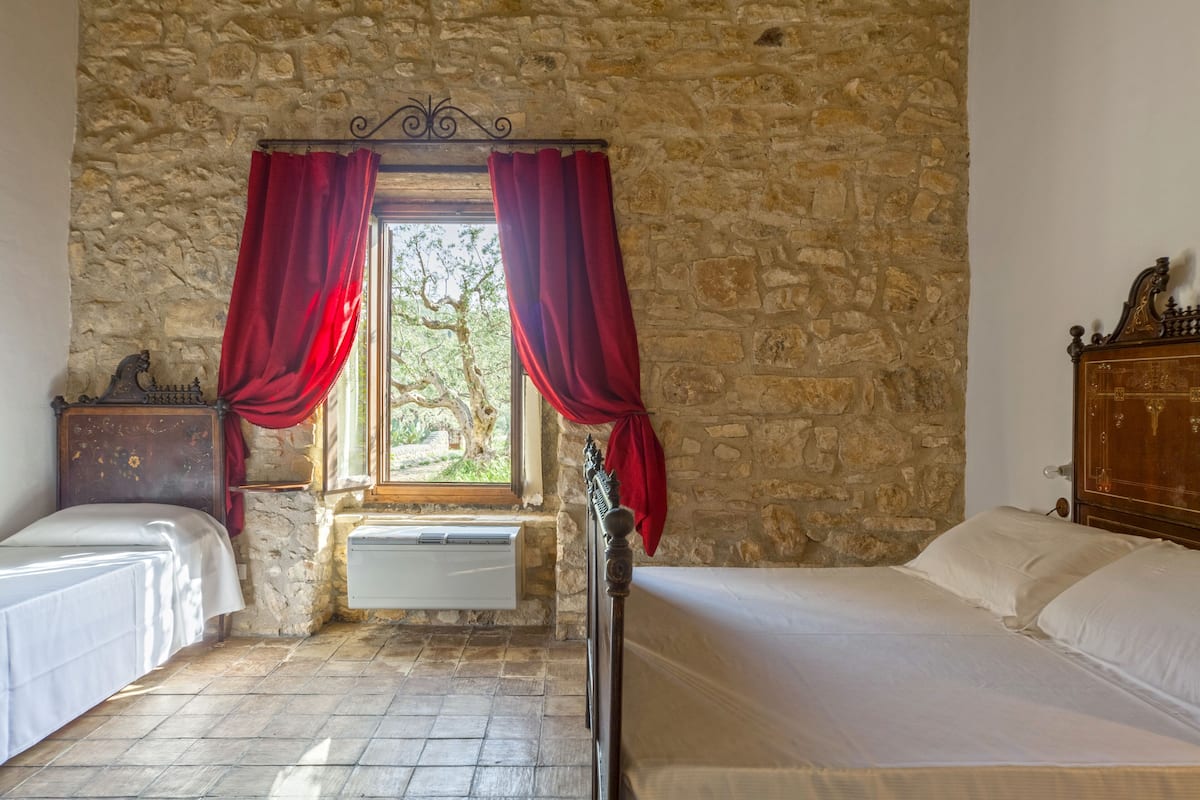 A room with stone walls, two beds, a window with red curtains, and a wooden dresser.