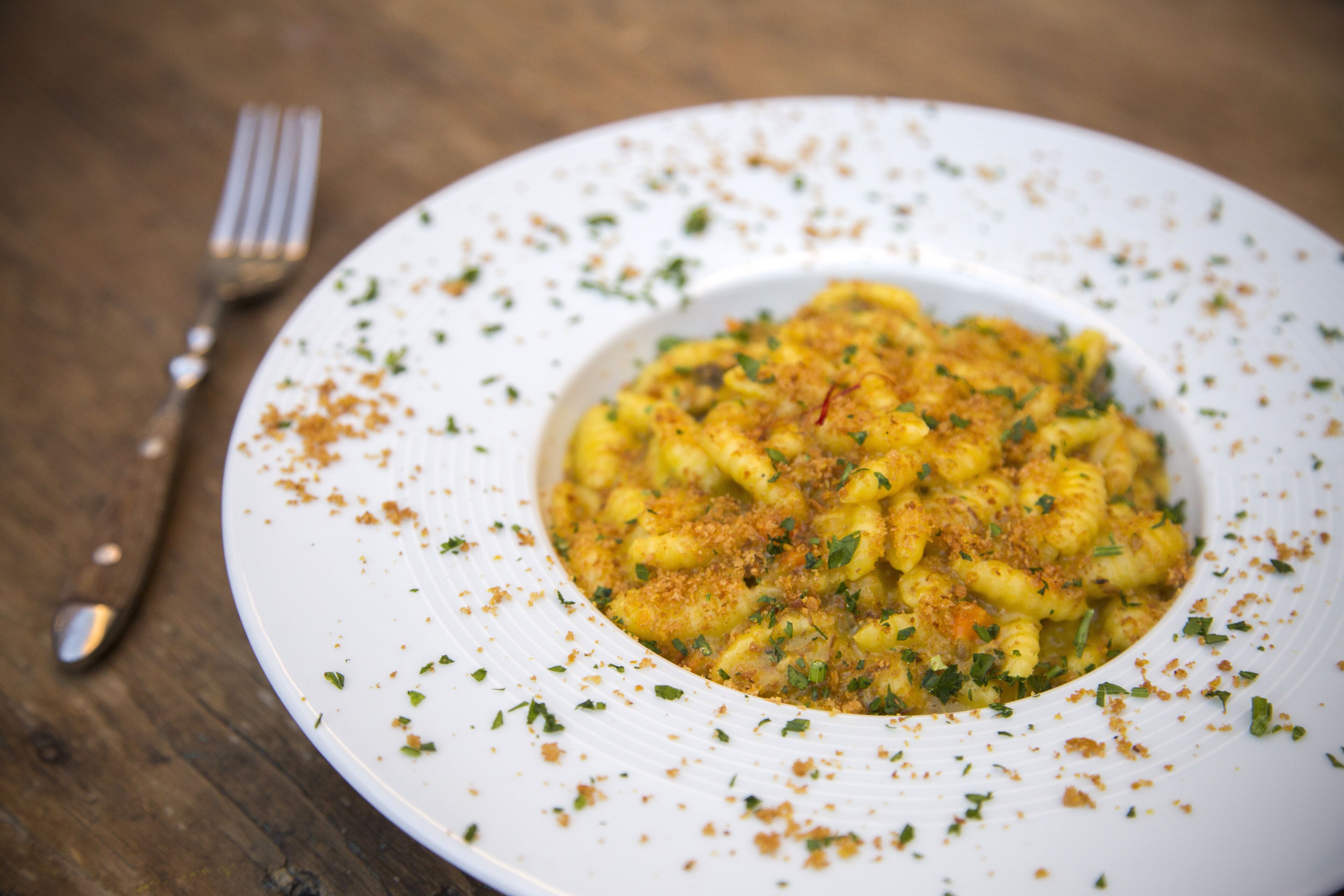 A white plate with pasta, garnished with herbs and breadcrumbs, on a wooden table.