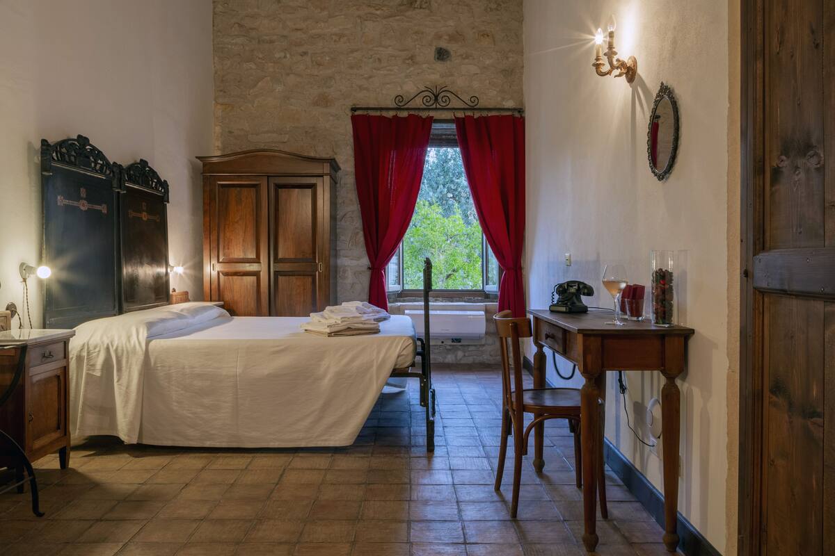 A room with a bed, a wooden desk, a chair, a wardrobe, and a window with red curtains.