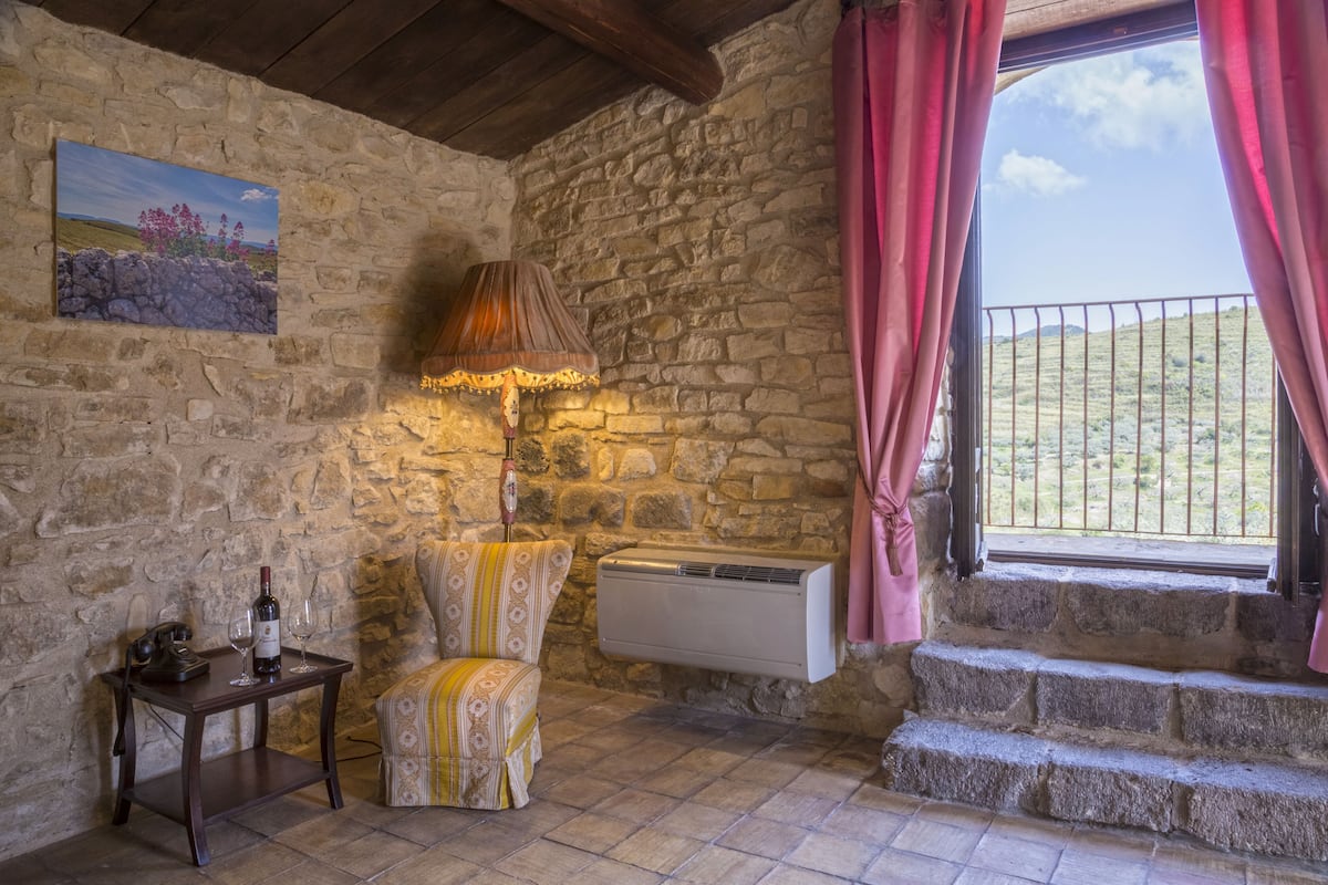 A rustic stone-walled room with a wooden table, a patterned armchair, a wall-mounted air conditioner, and a window with pink curtains.