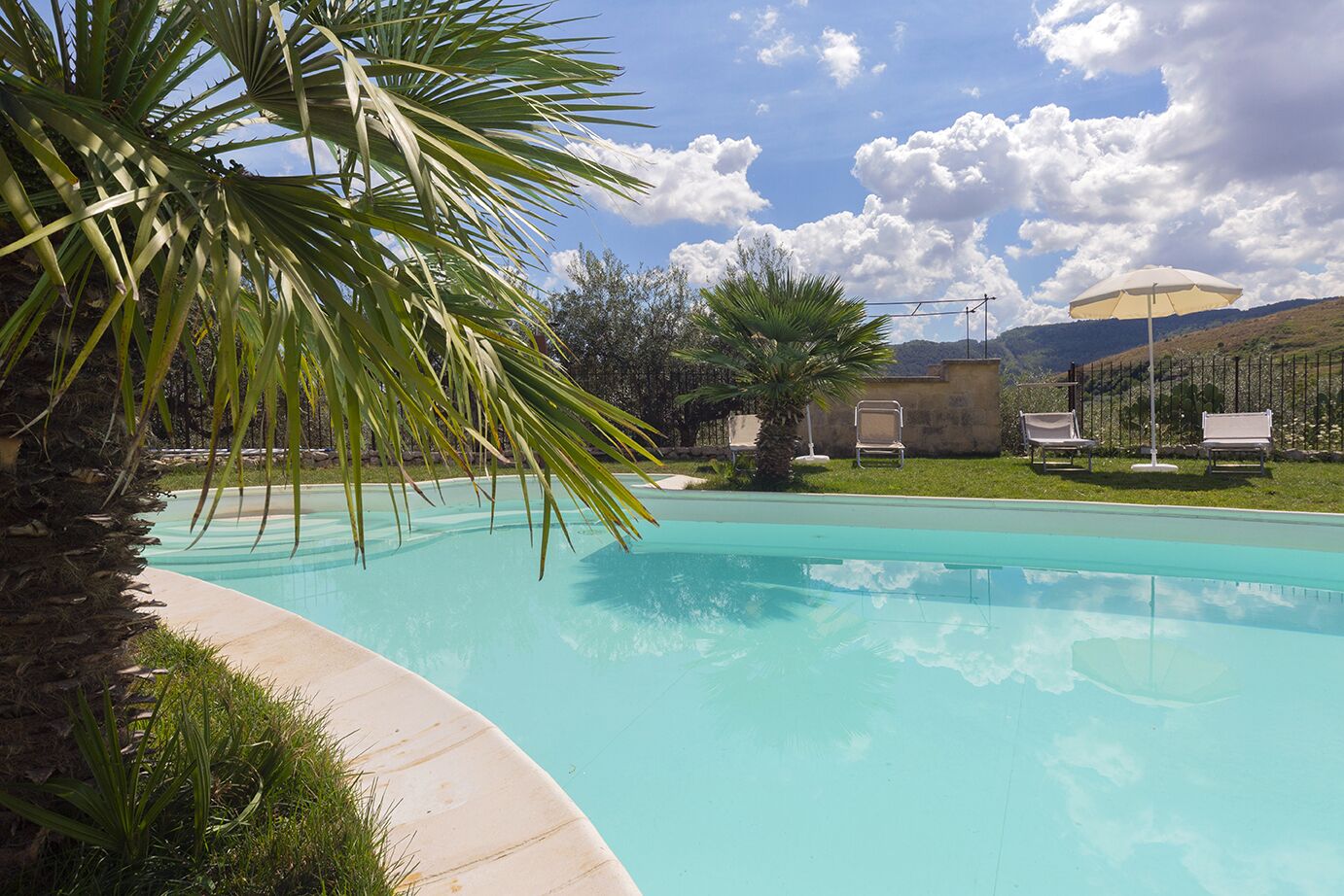 Une piscine d’eau cristalline, entourée de palmiers, et un petit bâtiment avec une clôture en arrière-plan.