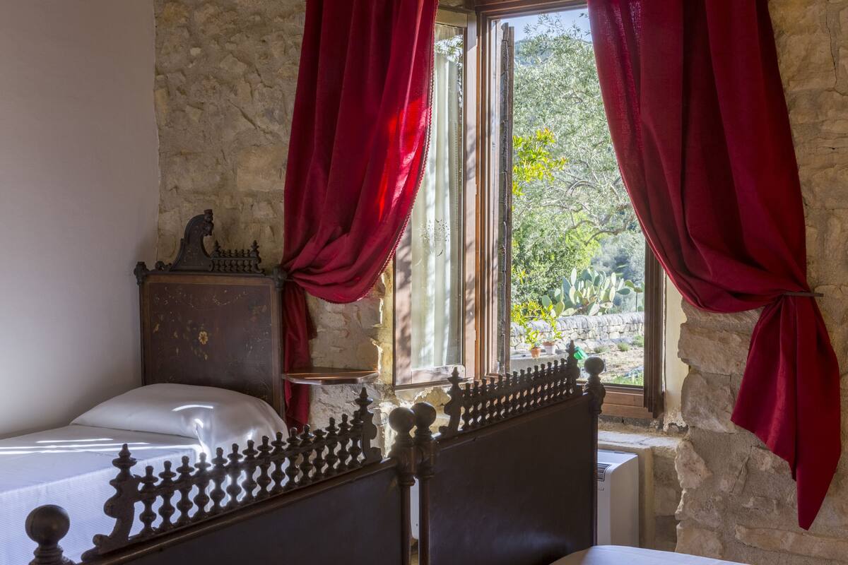 A bedroom with a stone wall, a wooden headboard, a bed with a white mattress, and red curtains.