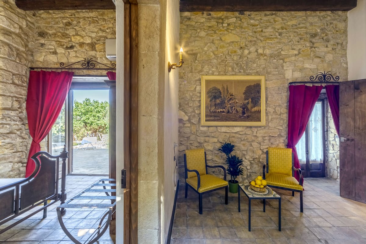A stone-walled room with a bed, two chairs, a table with fruit, and a painting on the wall.