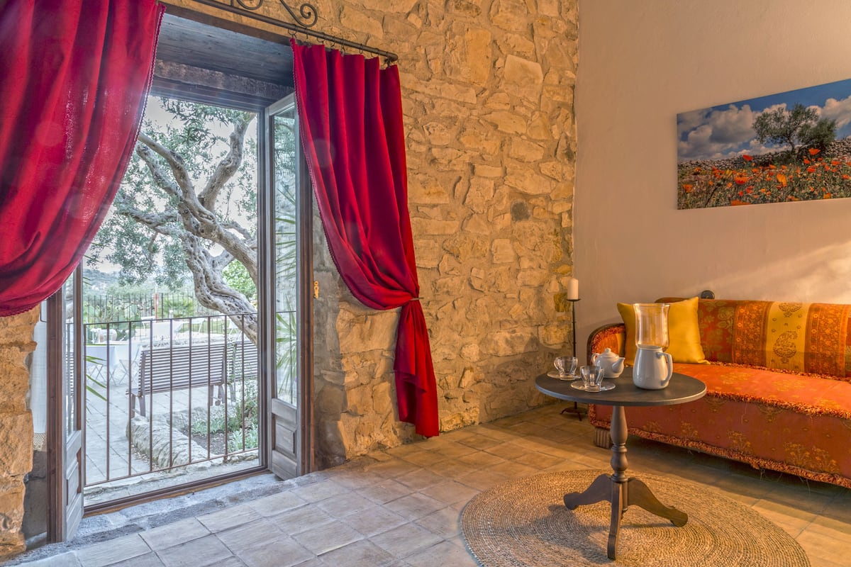 A room with a stone wall, a glass door with red curtains, a sofa with patterned cushions, and a painting of a landscape.