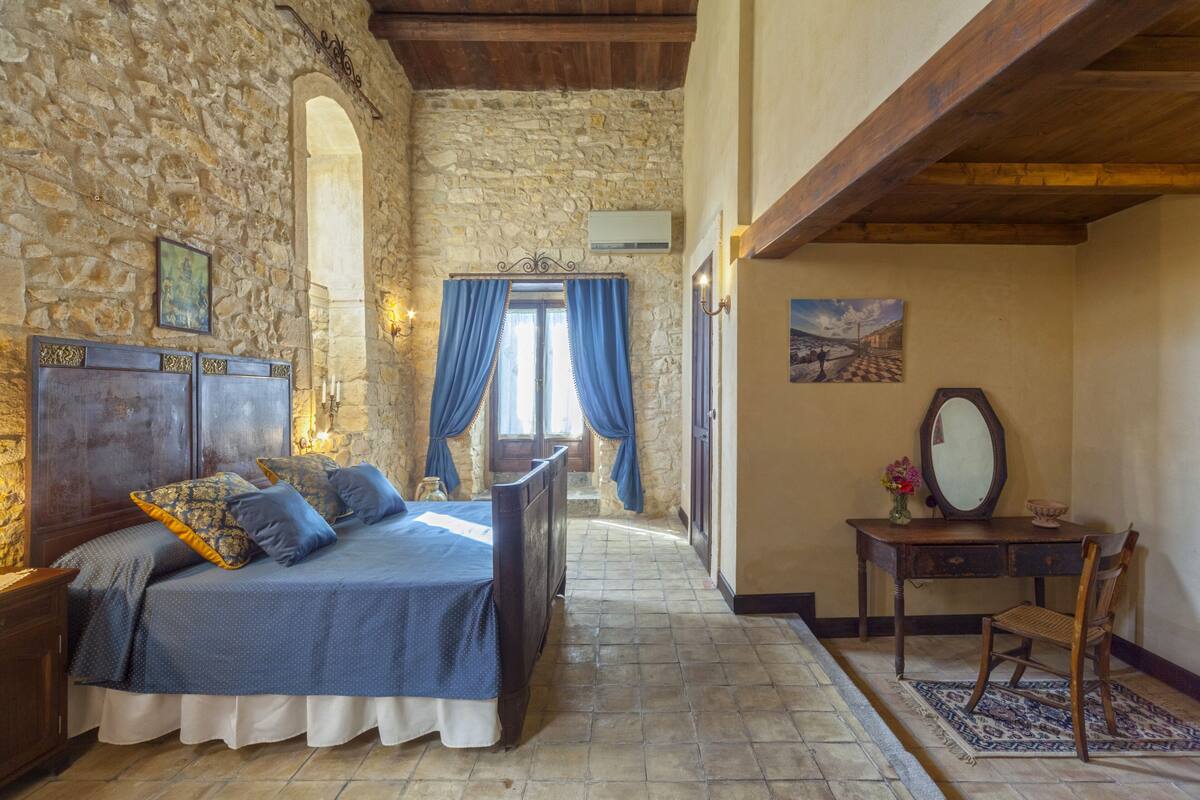 A bedroom with a stone wall, a wooden bed, a desk with a mirror, and a view through a window with blue curtains.
