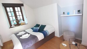 A compact bedroom with a bed, a small round table, a window, and a shelf with decorative items.