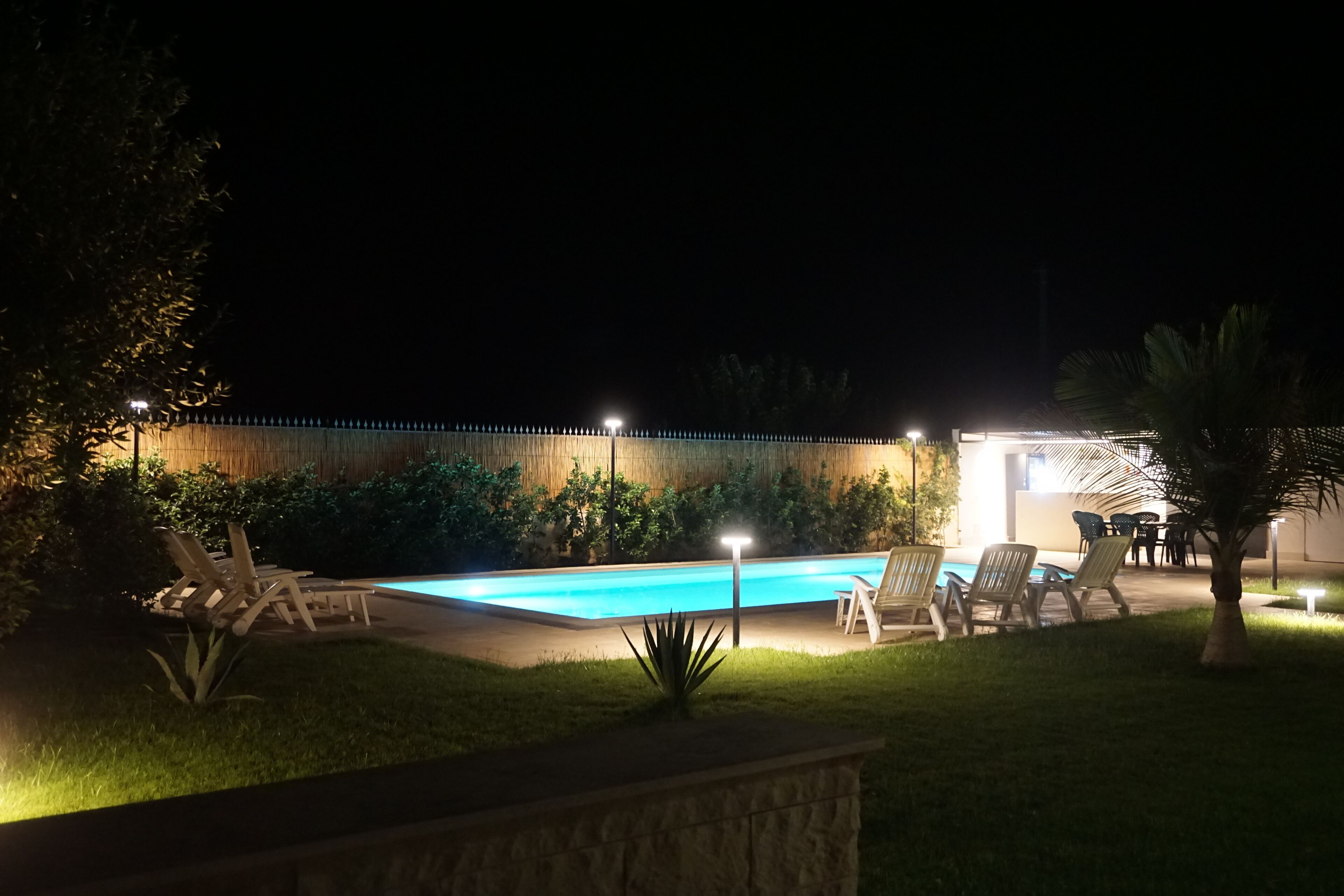 Zona de piscina a l'aire lliure de nit, amb gandules i una paret de pedra.