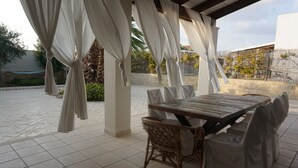 Eine Terrasse im Freien mit Holztisch und Korbstühlen, beschattet durch weiße Vorhänge.