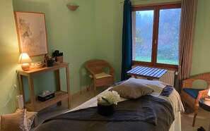 Ein Zimmer mit einem Bett, einem kleinen Tisch mit Lampe, einem Korbstuhl und einem Fenster mit Vorhängen.