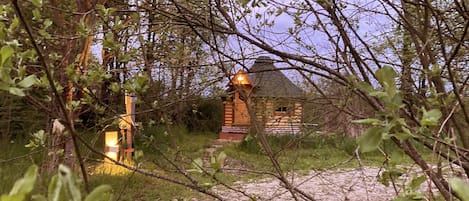 A rustic wooden cabin with a thatched roof, surrounded by dense foliage and a gravel path leading to it.