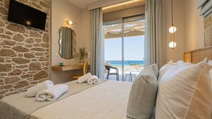 A hotel room with a stone wall, a flat-screen TV, a large bed with white linens, a view of the beach, and a small table with chairs. - New Villa★Prive Pool★Walk to all & Falasarna beach★FREE Daily Breakfast (Falasarna Crete)