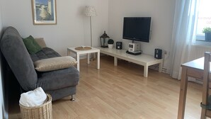 Ein Wohnzimmer mit grauem Sofa, einem kleinen weißen Tisch, einem Fernseher und einer Lampe.