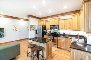 A kitchen with wooden cabinets, a stainless steel refrigerator, a microwave, a stove, and a sink. There are two stools and a green couch.