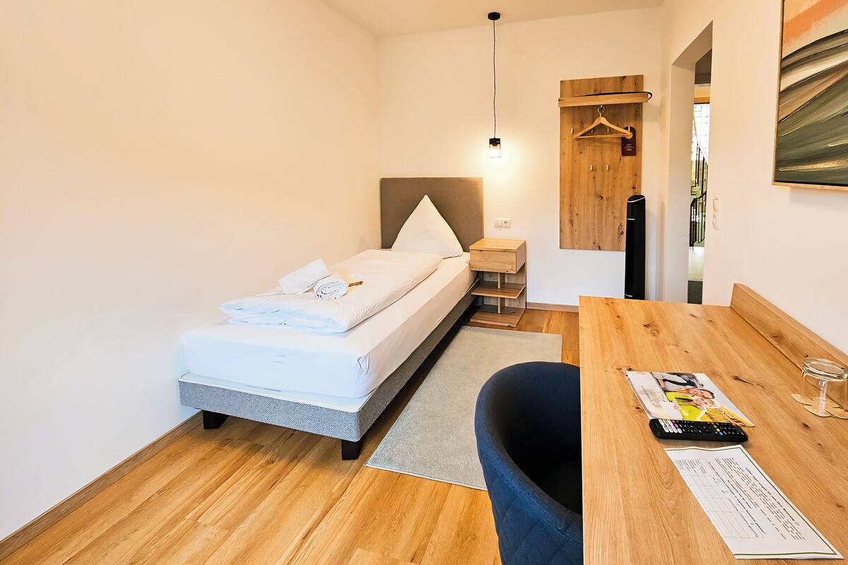 A modern hotel room with a bed, a wooden desk, a chair, a small table, a hanging light, and a wooden door.