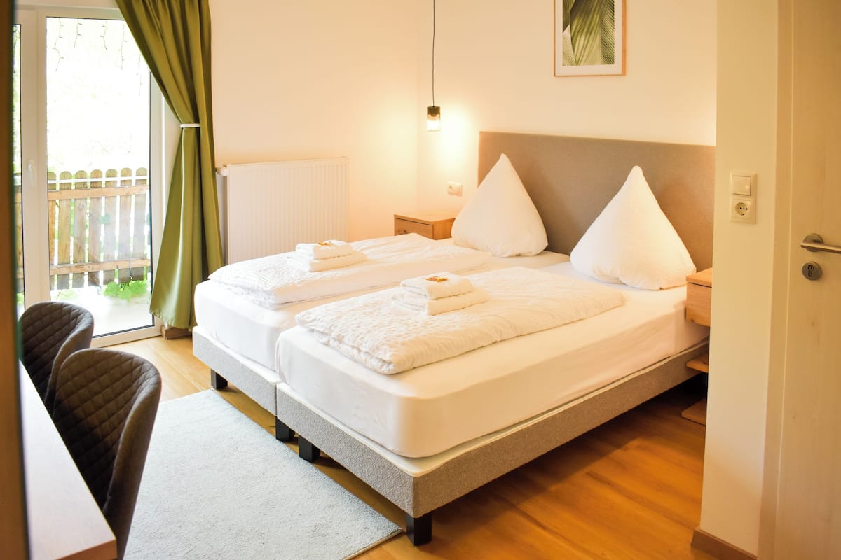 A double bed with two pillows, a bedside table, a chair, and a view of a wooden deck through a window with curtains.