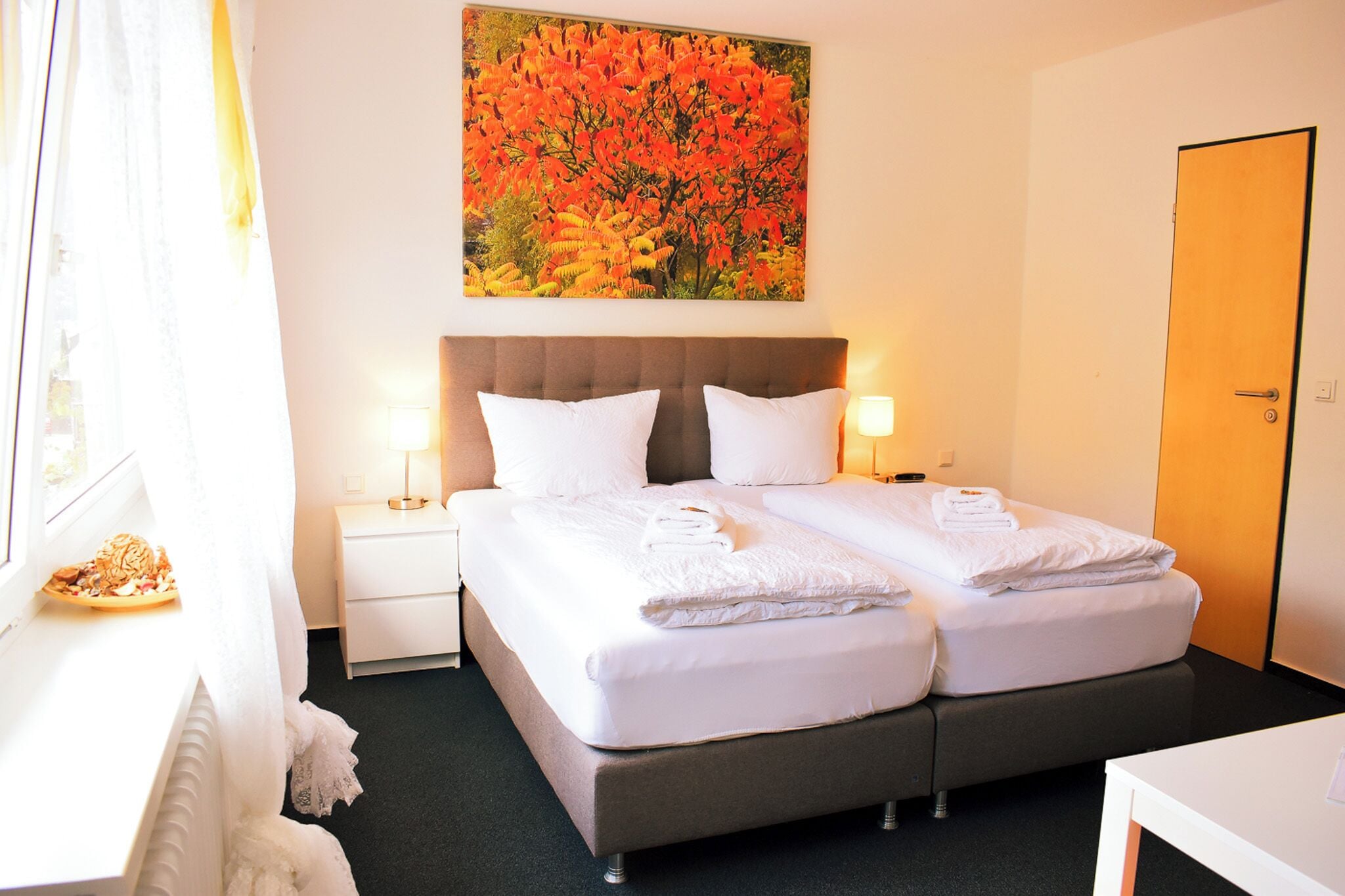 A double bed with white linens, two bedside tables with lamps, a large painting of autumn foliage, and a door to the right.