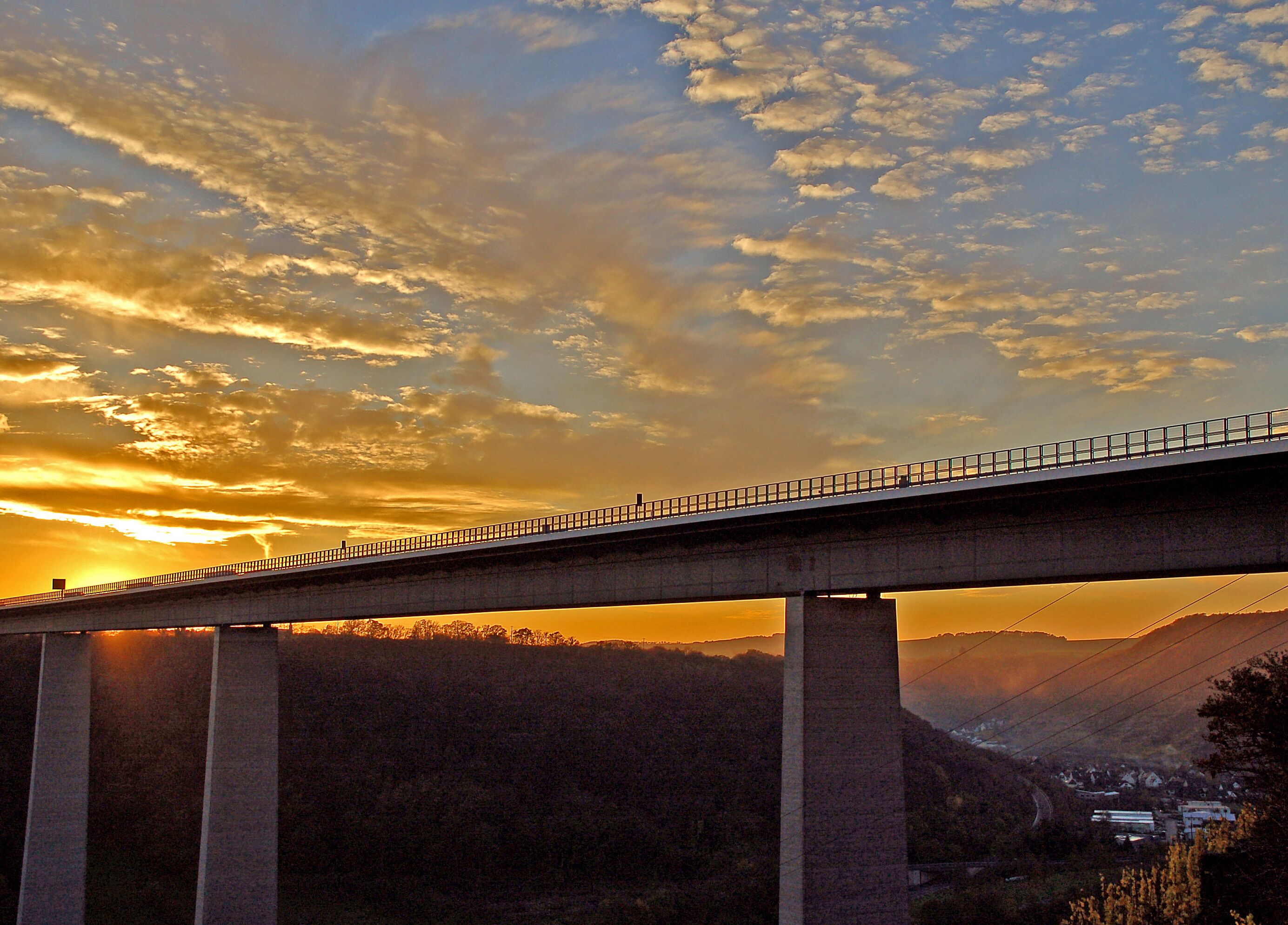 Un ponte con percorso pedonale e parapetti, che si estende su una valle con un tramonto sullo sfondo.