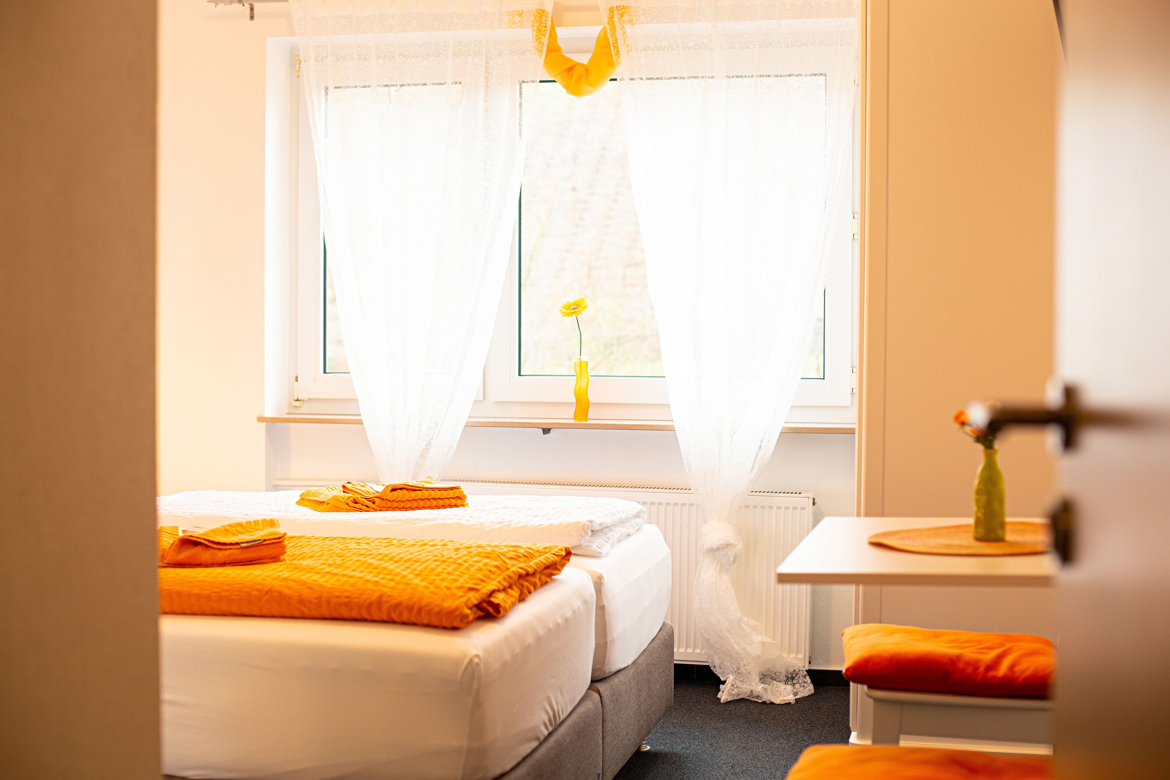 A hotel room with two beds, a window with curtains, a radiator, and a small table with a vase.
