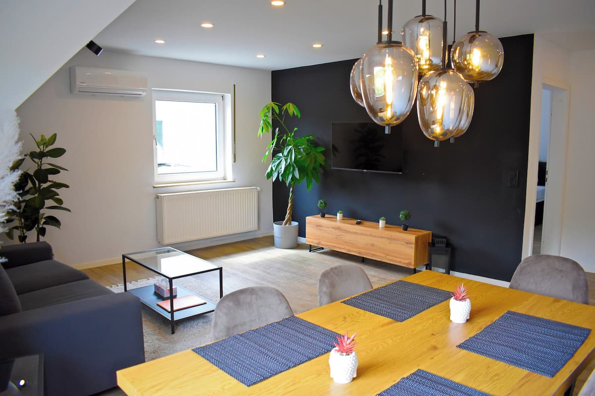 A modern living room with a dining area, featuring a sofa, a glass coffee table, a wooden dining table with placemats, and a hanging light fixture.