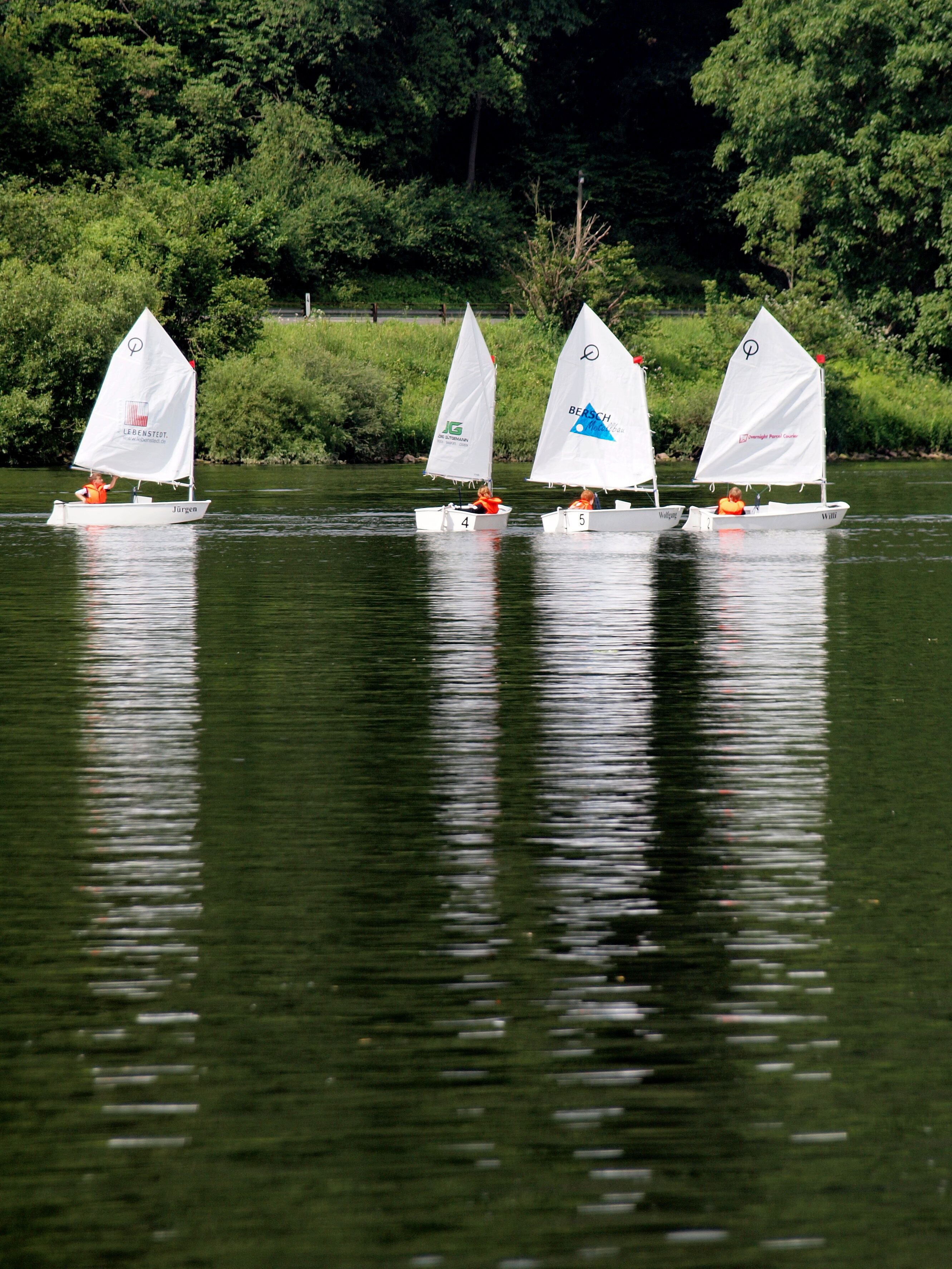 Four sailboats on a calm river with lush greenery in the background.