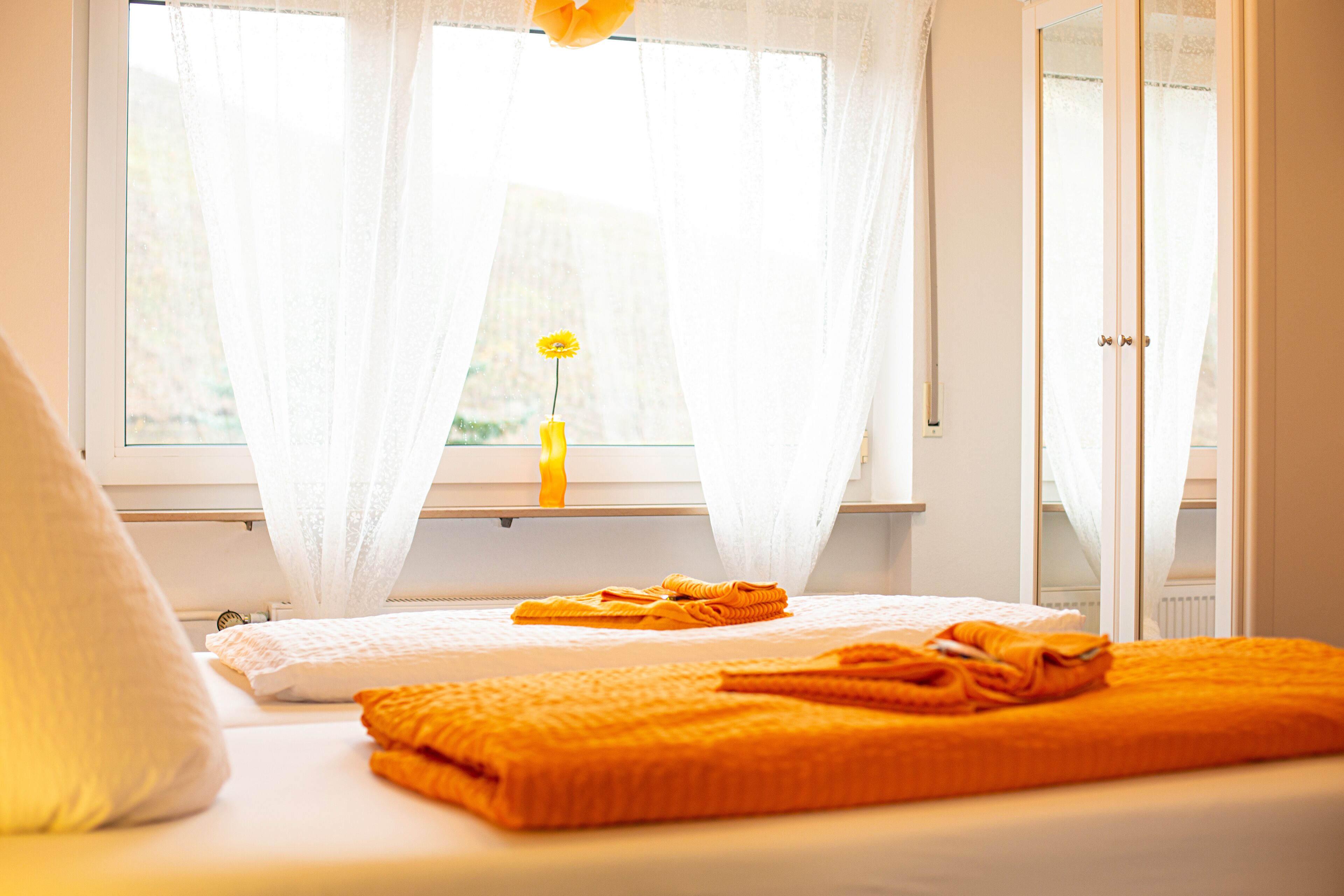 Un dormitorio con una cama, una ventana con cortinas translúcidas y un florero con una flor.