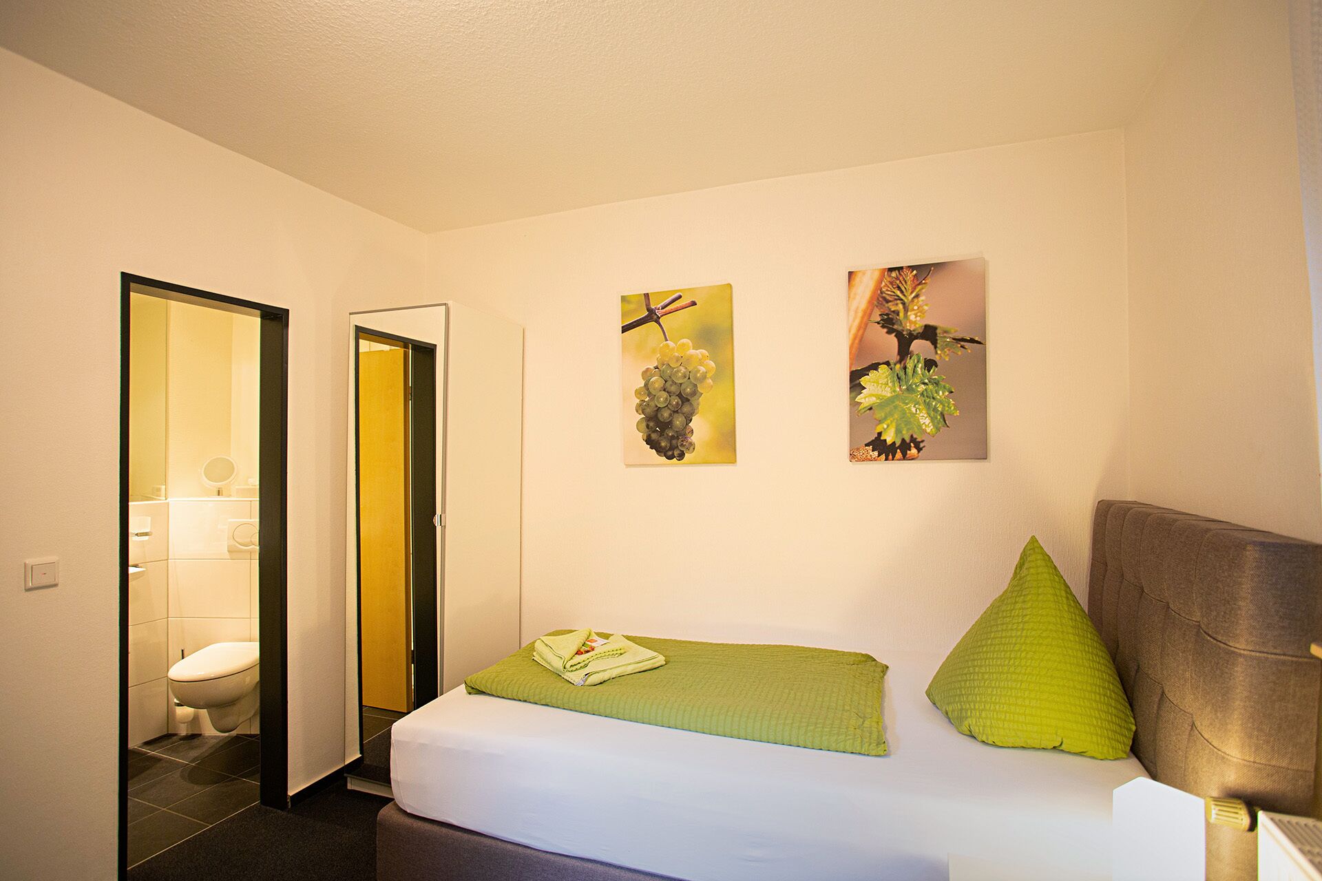A hotel room with a bed, a green pillow, a green blanket, and two wall art pieces depicting plants.