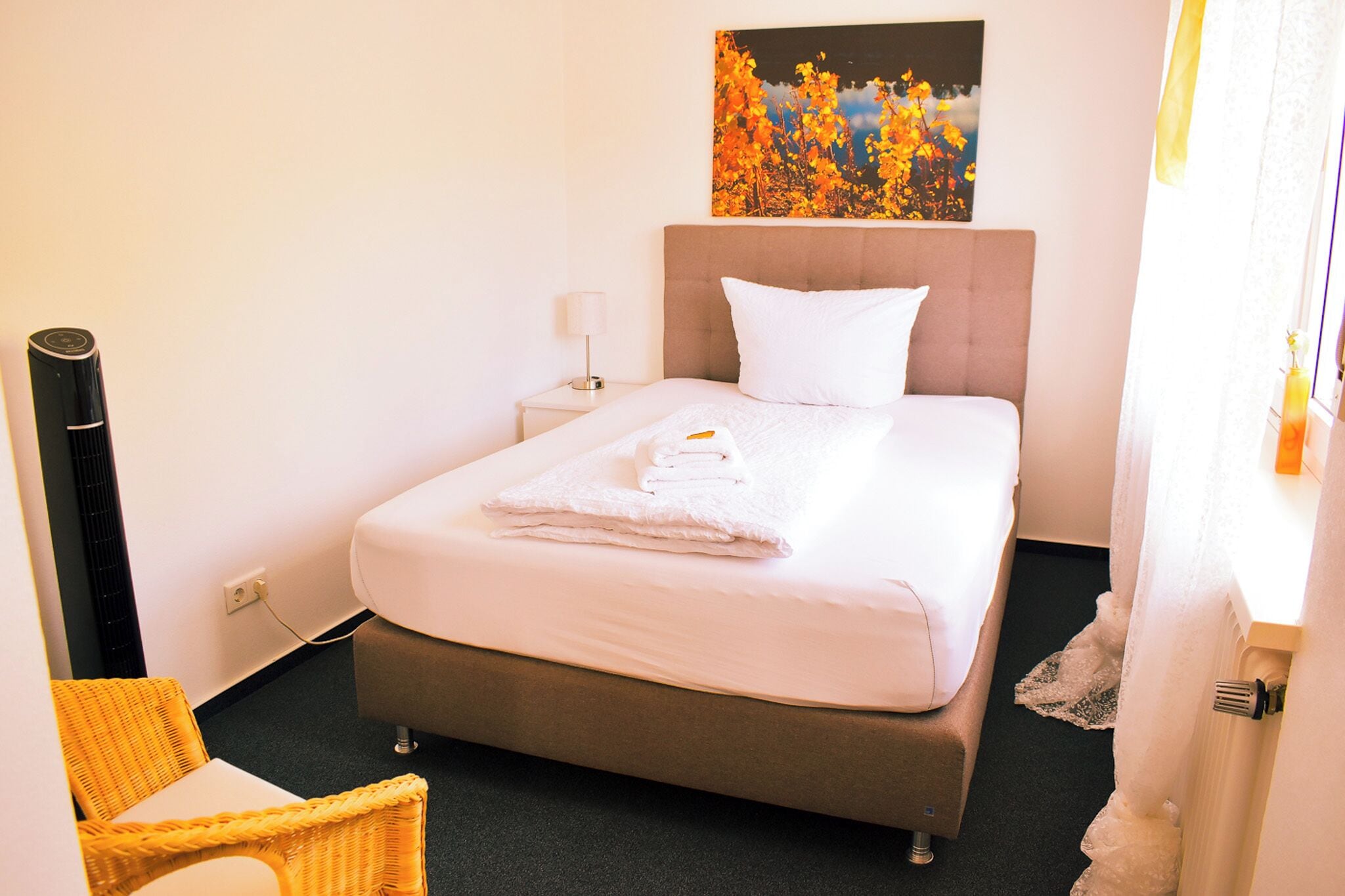 Una cama bien hecha con edredón blanco y almohadas, una lámpara de noche y un cuadro de hojas otoñales en la pared.
