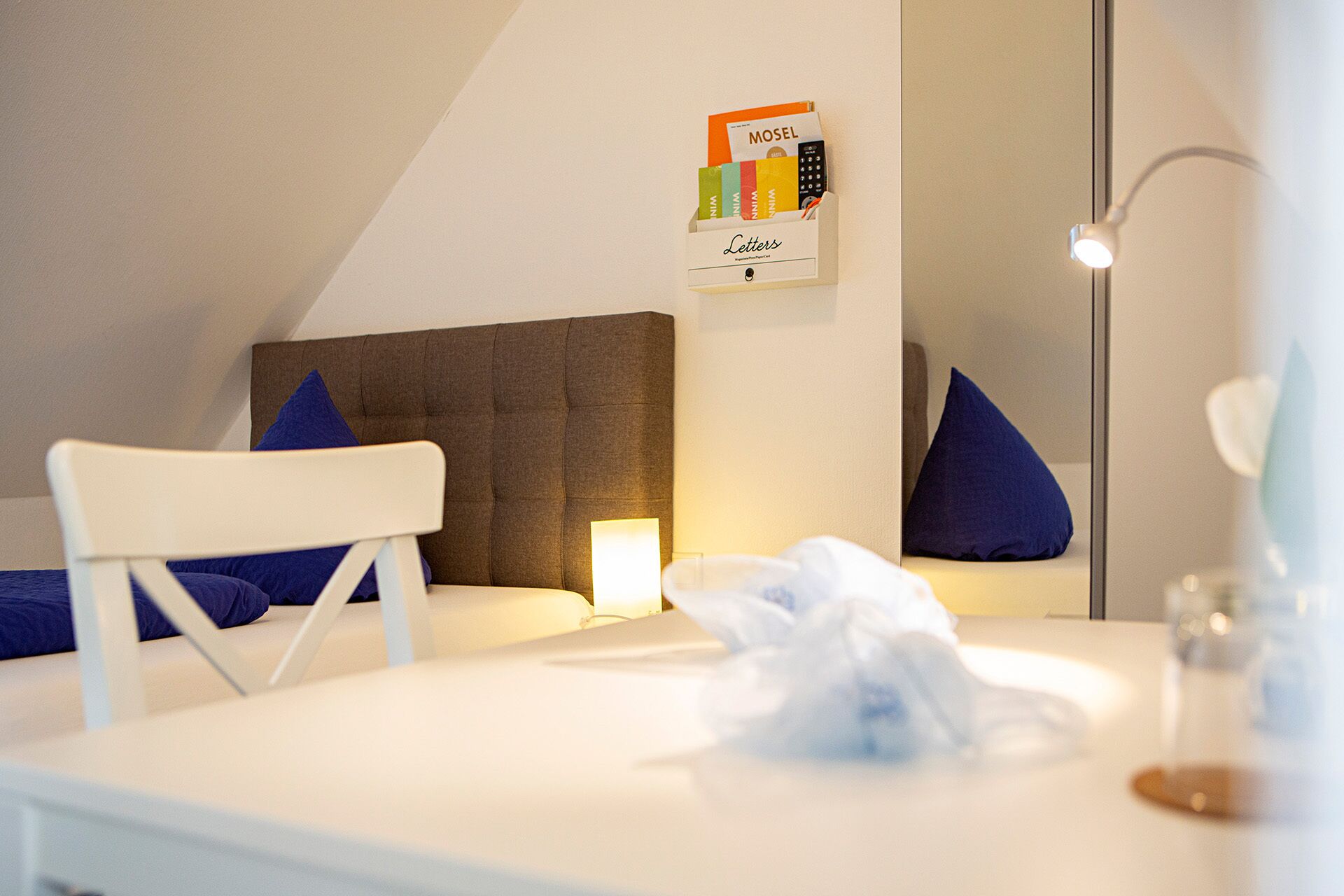 A neatly arranged hotel room with a bed, a bedside table, a chair, a bedside lamp, a mirror, and a wall-mounted shelf with a book and a remote control.