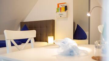 A neatly arranged hotel room with a bed, a bedside table, a chair, a bedside lamp, a mirror, and a wall-mounted shelf with a book and a remote control.