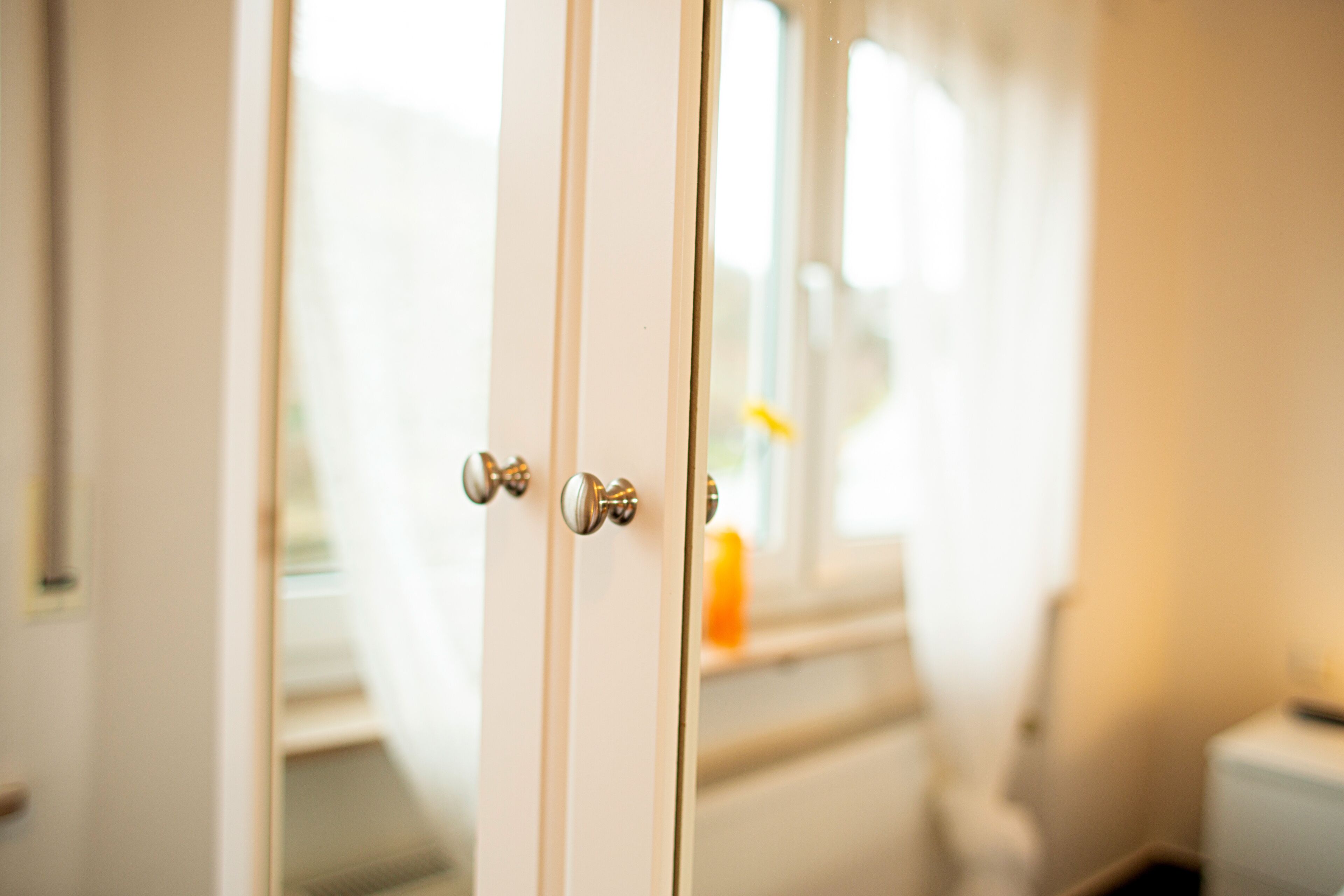 Primer plano de una puerta blanca con manijas plateadas, que da paso a un cuarto con ventana y un área de baño visible.