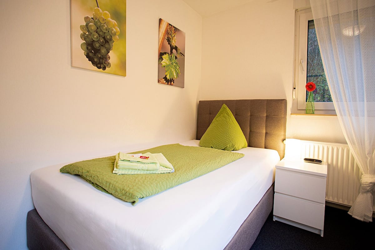 A neatly made bed with a green blanket and pillows, a bedside table with a remote control, and a window with a view of trees.
